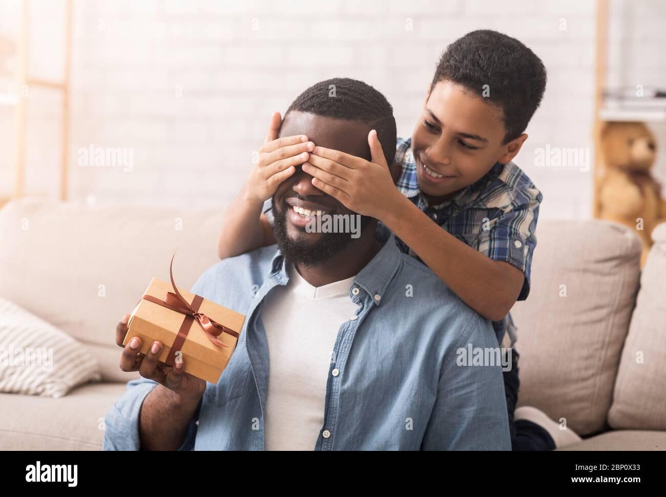 Happy son congratulating his afro dad, covering eyes and giving present Stock Photo
