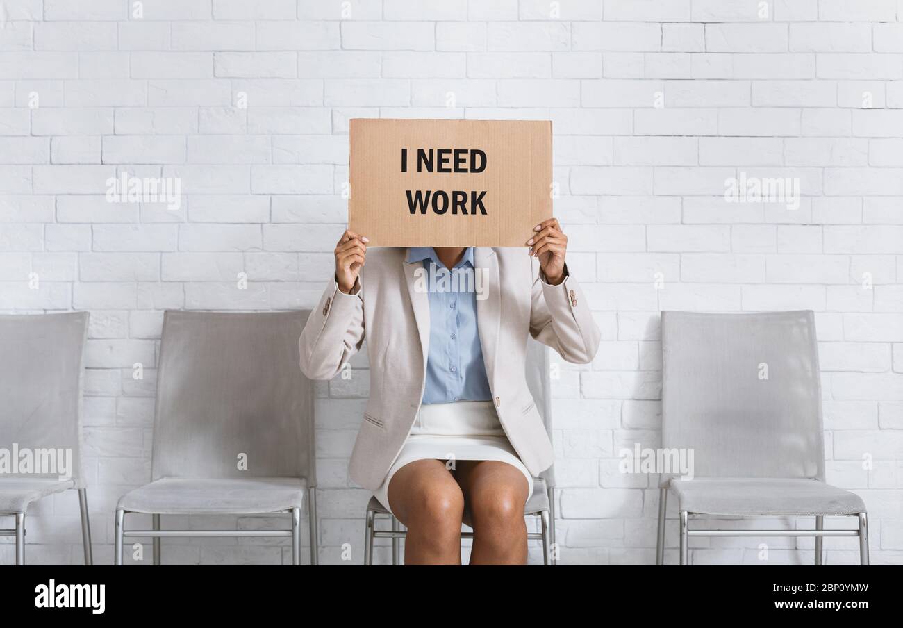 Black woman in business wear hiding behind sign with inscription I NEED WORK while waiting for work interview in office Stock Photo