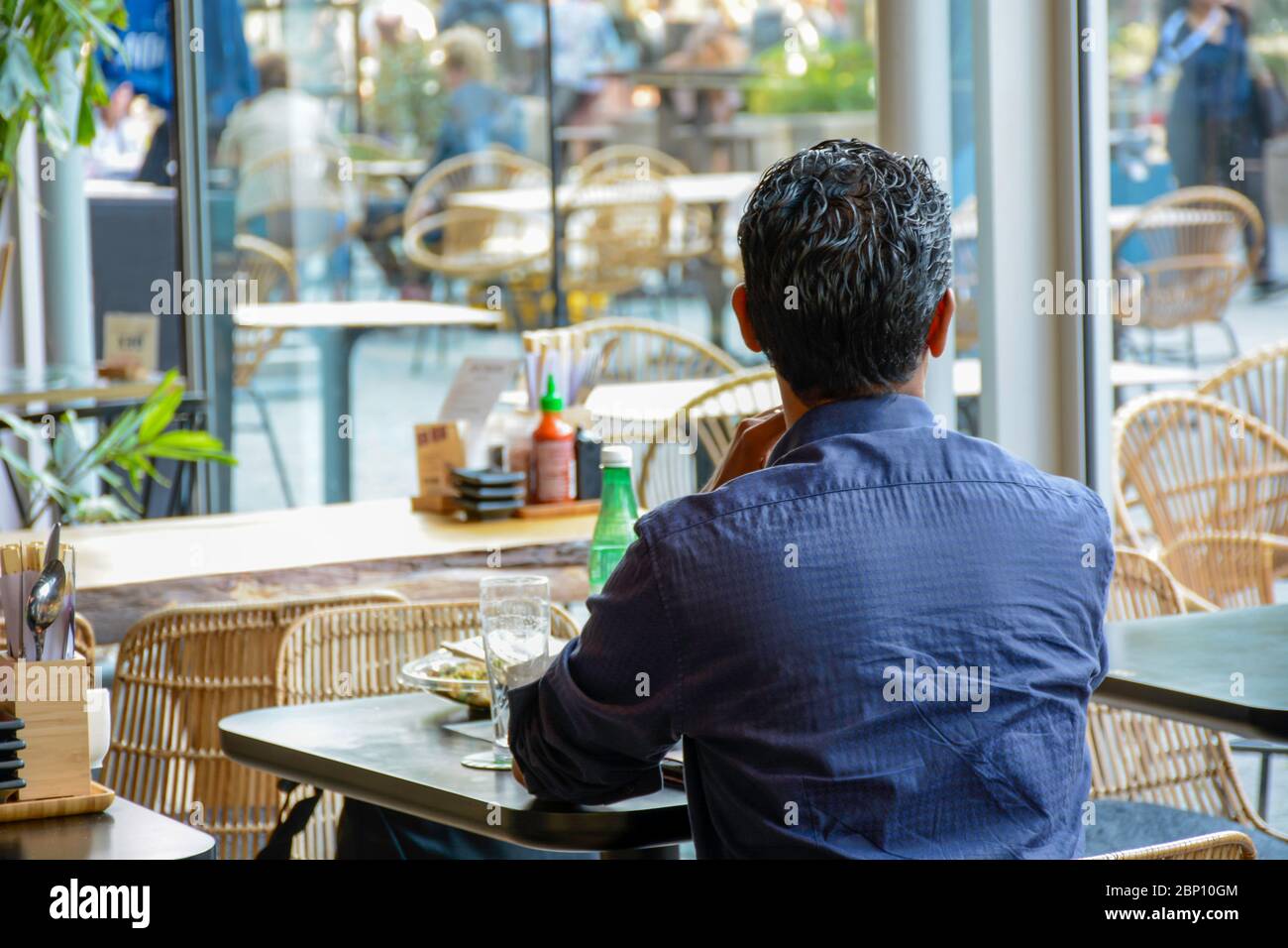 Man from behind sits alone in a modern cafe restaurant and looks outside. He is waiting for his companion to return. There is no one else around him. Stock Photo