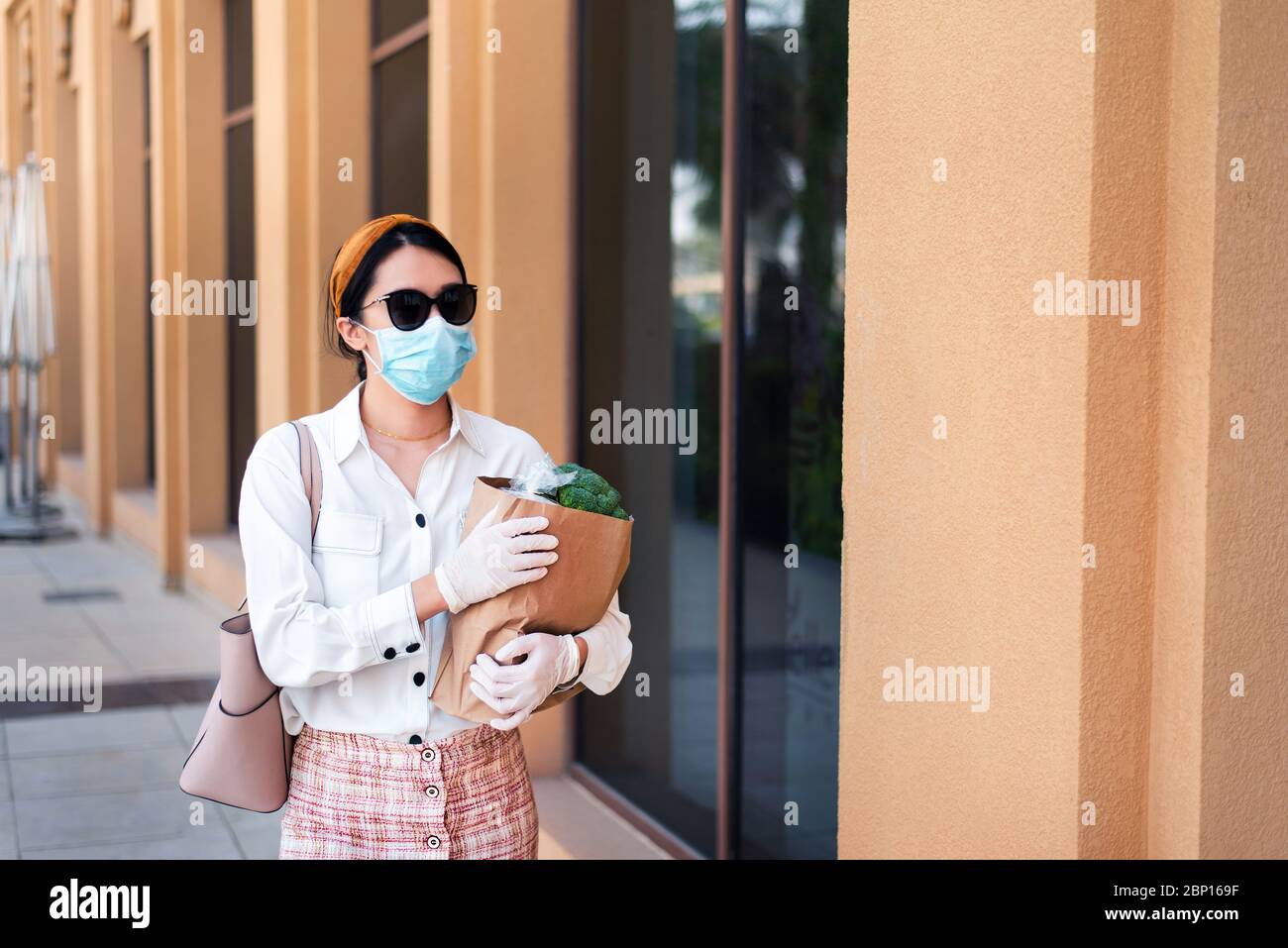 Asian woman carrying shopping bag with groceries while wearing mask and gloves to prevent virus spread during coronavirus pandemic Stock Photo