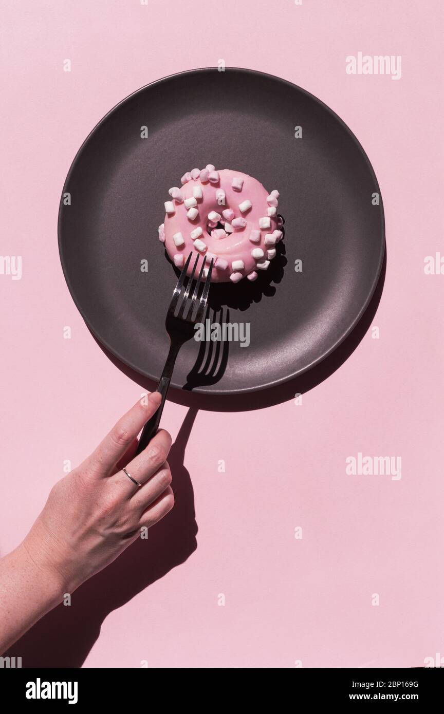 Light pink delicious donat with white marshmallows at the dark grey ceramic plate on the rosy background. Hand of woman with ring with freckles is Stock Photo
