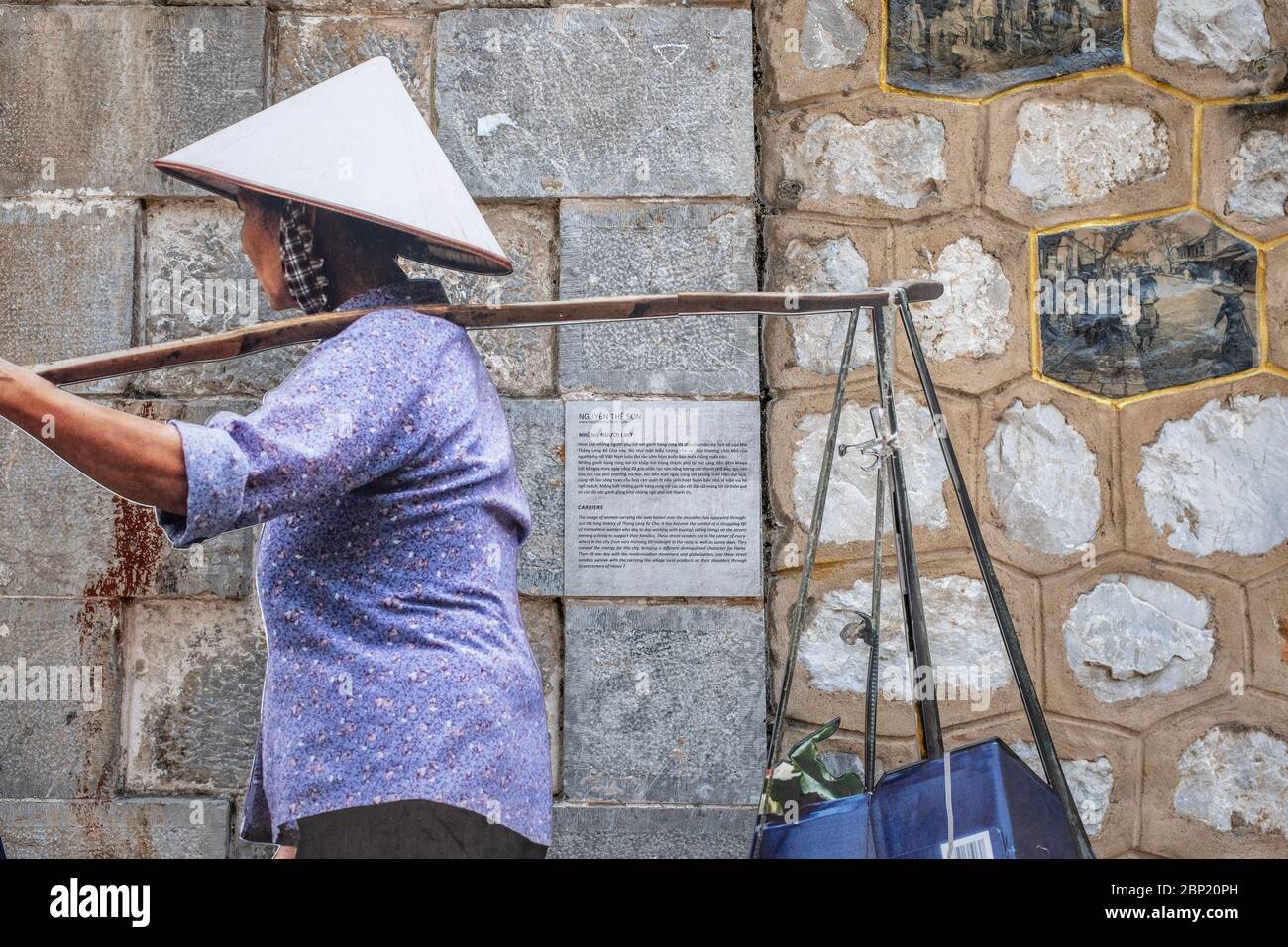 Hanoi, Vietnam - May 1, 2018: Woman carrying yoke baskets, wearing traditional stray hat, with stone wall in the background Stock Photo