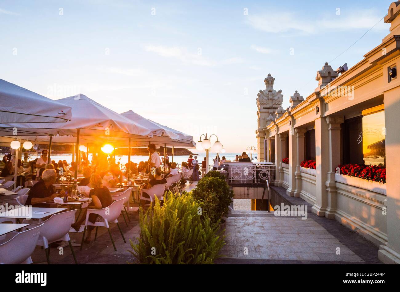 Donostia, Gipuzkoa, Basque Country, Spain - July 12th, 2019 : People relax at sunset in a chic sidewalk cafe in the promenade of La Concha beach. Stock Photo