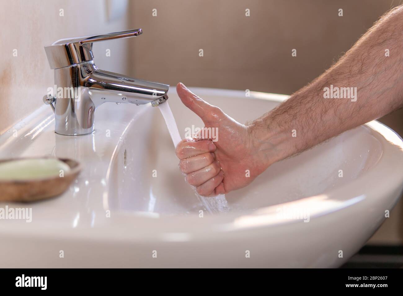 Hand of young caucasian man under a running water tap, showing thumb up gesture. Concept of personal hygiene, economical usage of water, saving money Stock Photo