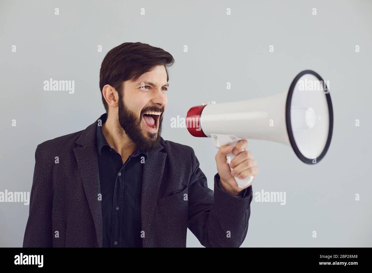 A man in a jacket shouts in a bullhorn on a gray background. Stock Photo
