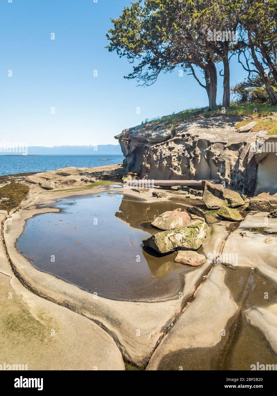 At low tide on a sunny day, a tidepool is framed by eroded channels in the smooth sandstone beach on an islet in British Columbia's Flat Top Islands. Stock Photo