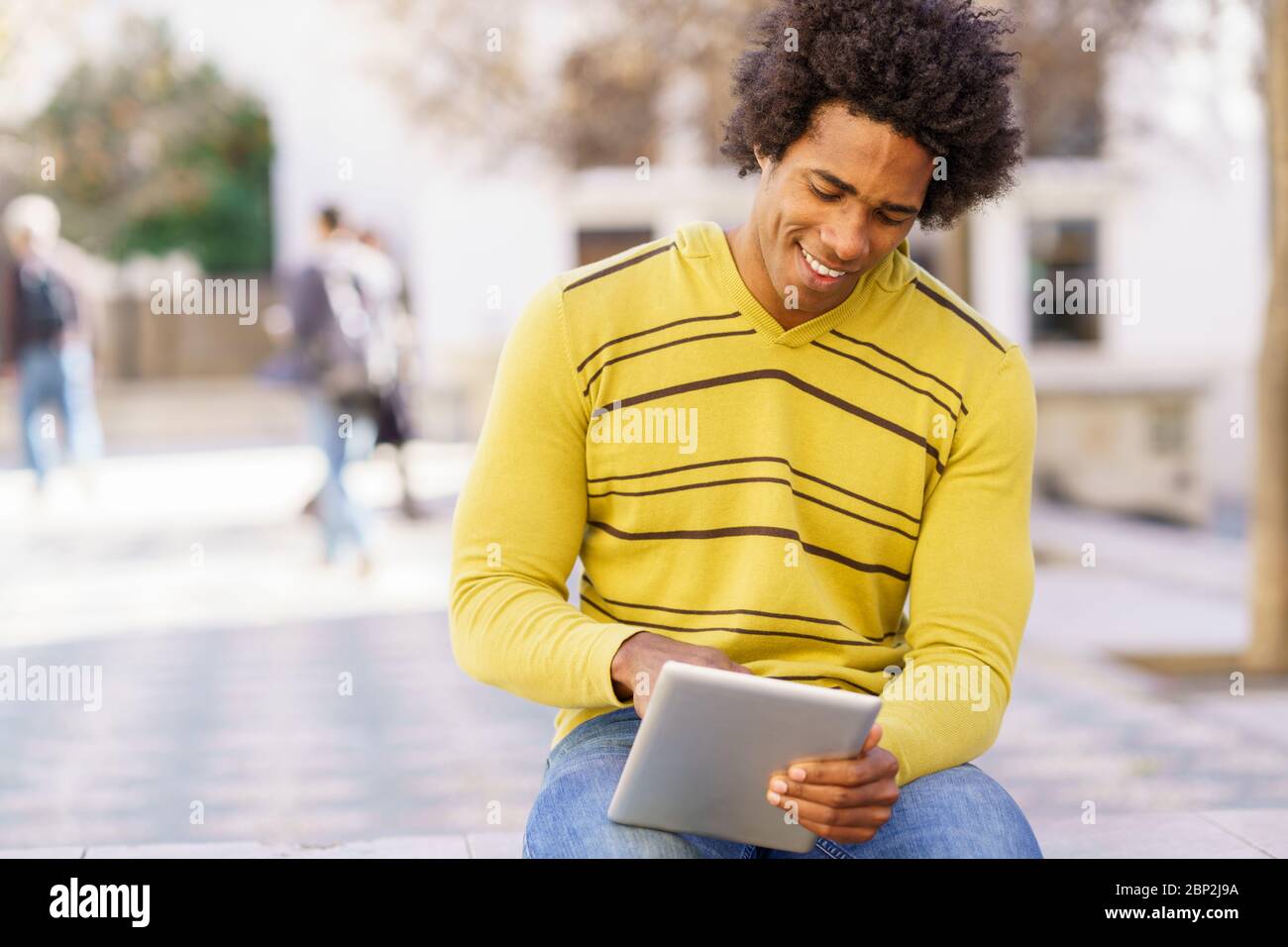 Black man using a digital tablet sitting on a bench outdoors. Stock Photo