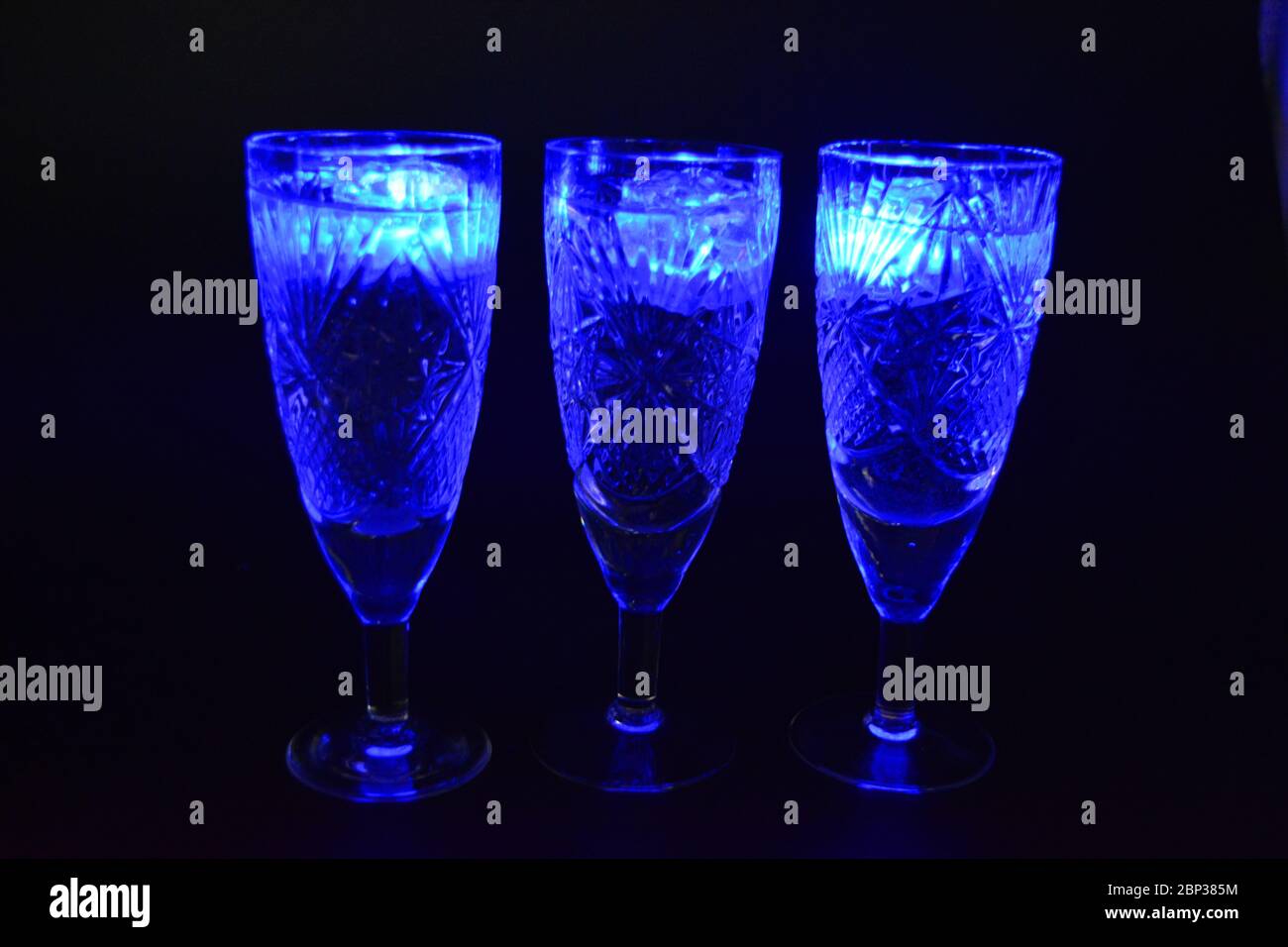 Crystal champagne glasses with carvings in the form of snowflakes are located on a black matte background. Bright multicolor doidic glowing ice cubes Stock Photo