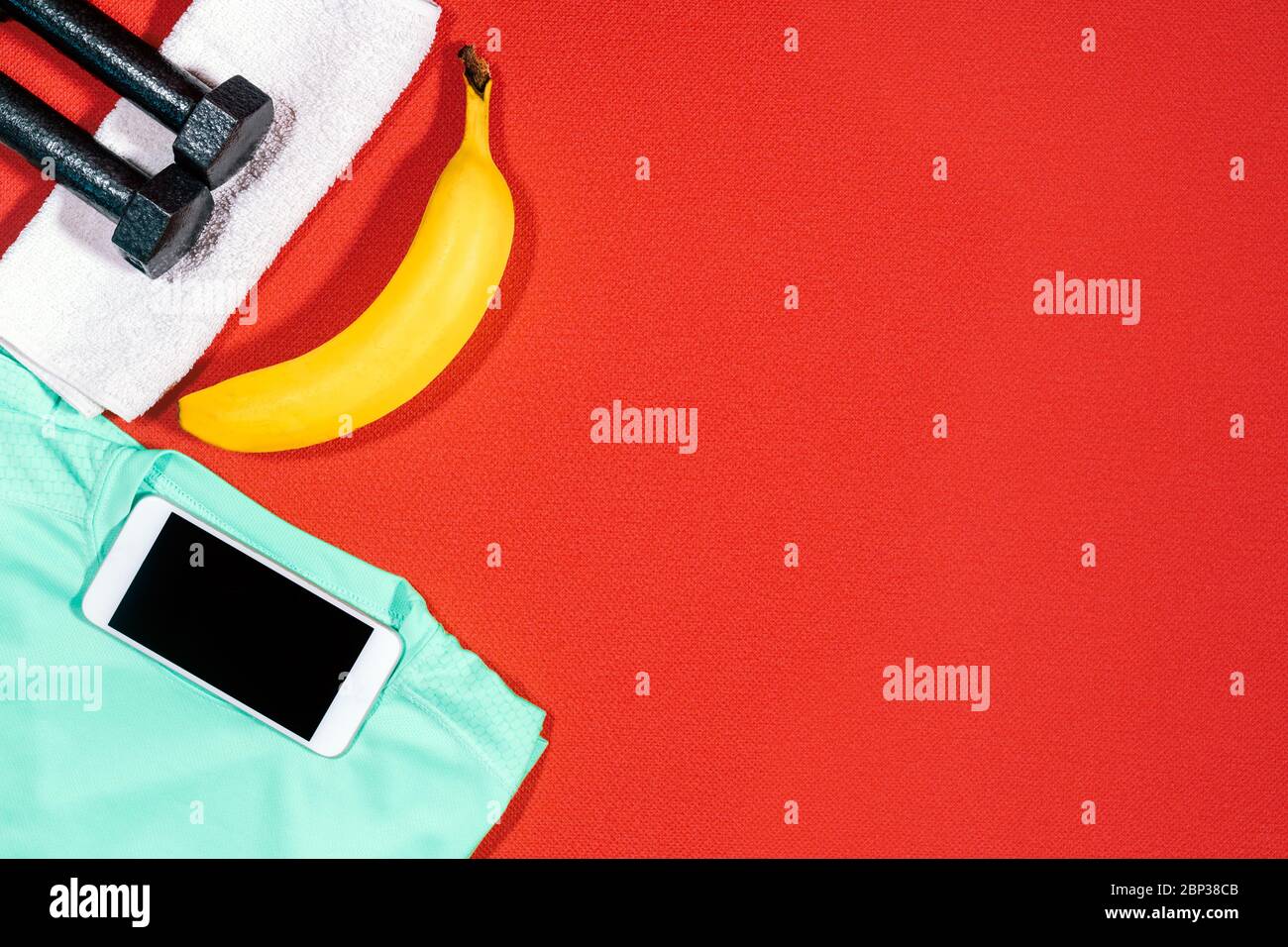 Set of sports attributes. Weights, a towel, a sports shirt, a mobile phone and a banana on a red mat for sport. Stock Photo