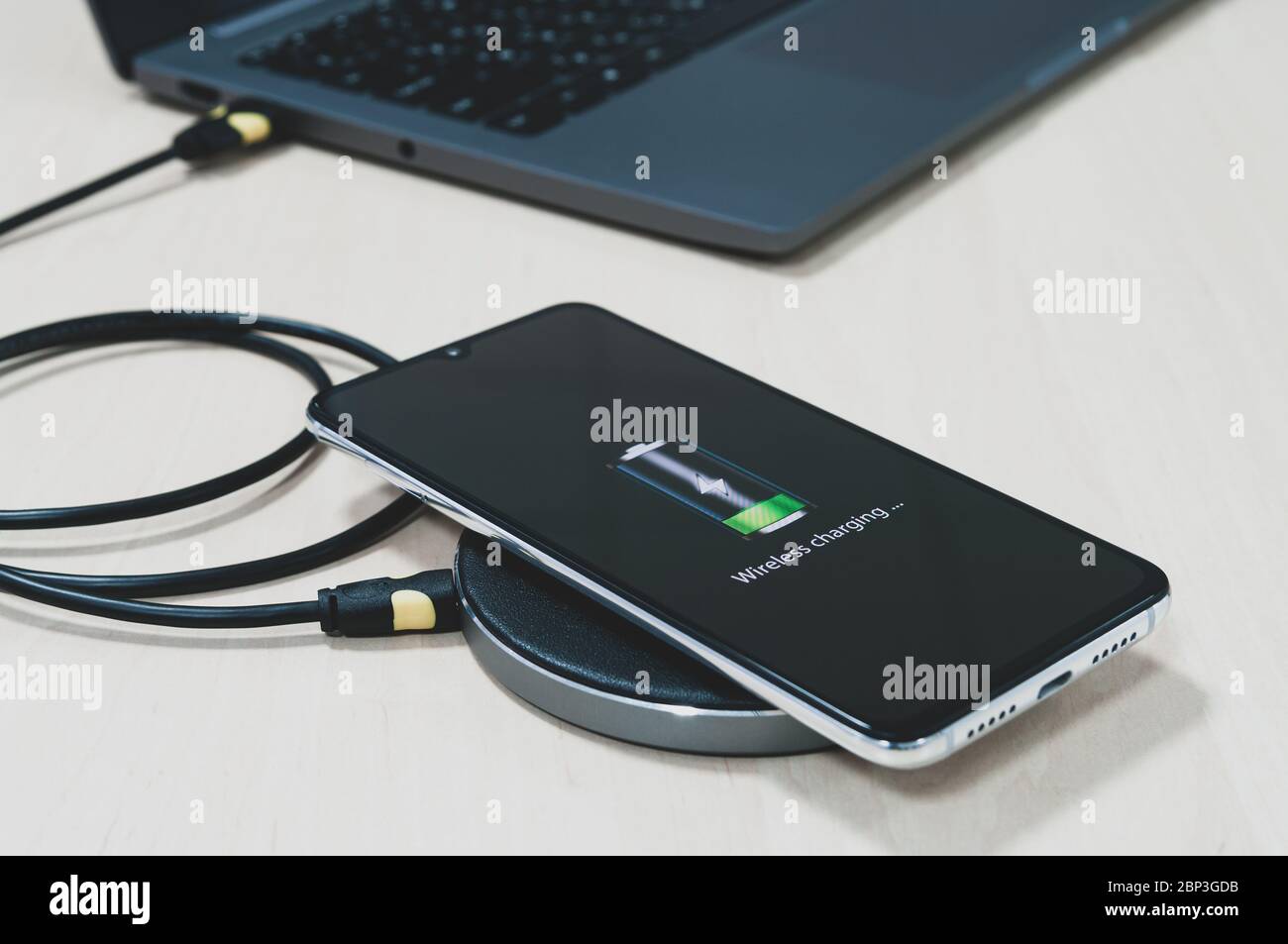 Modern smartphone are charging from wireless charger lies next to the laptop on a wooden table. Modern technology concept. Selective focus Stock Photo