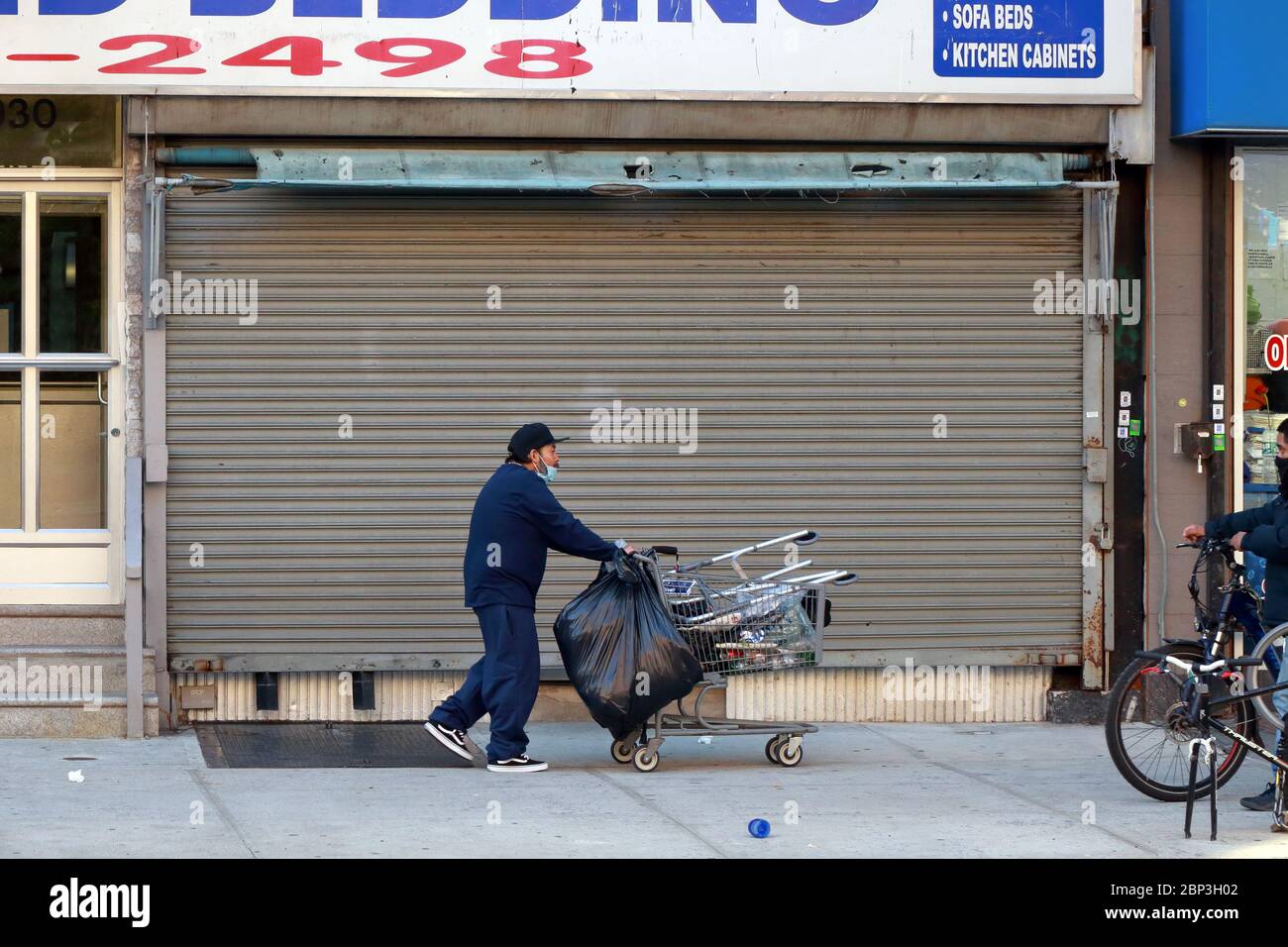 A person with a shopping cart against a shuttered storefront roll down gate in New York City Stock Photo