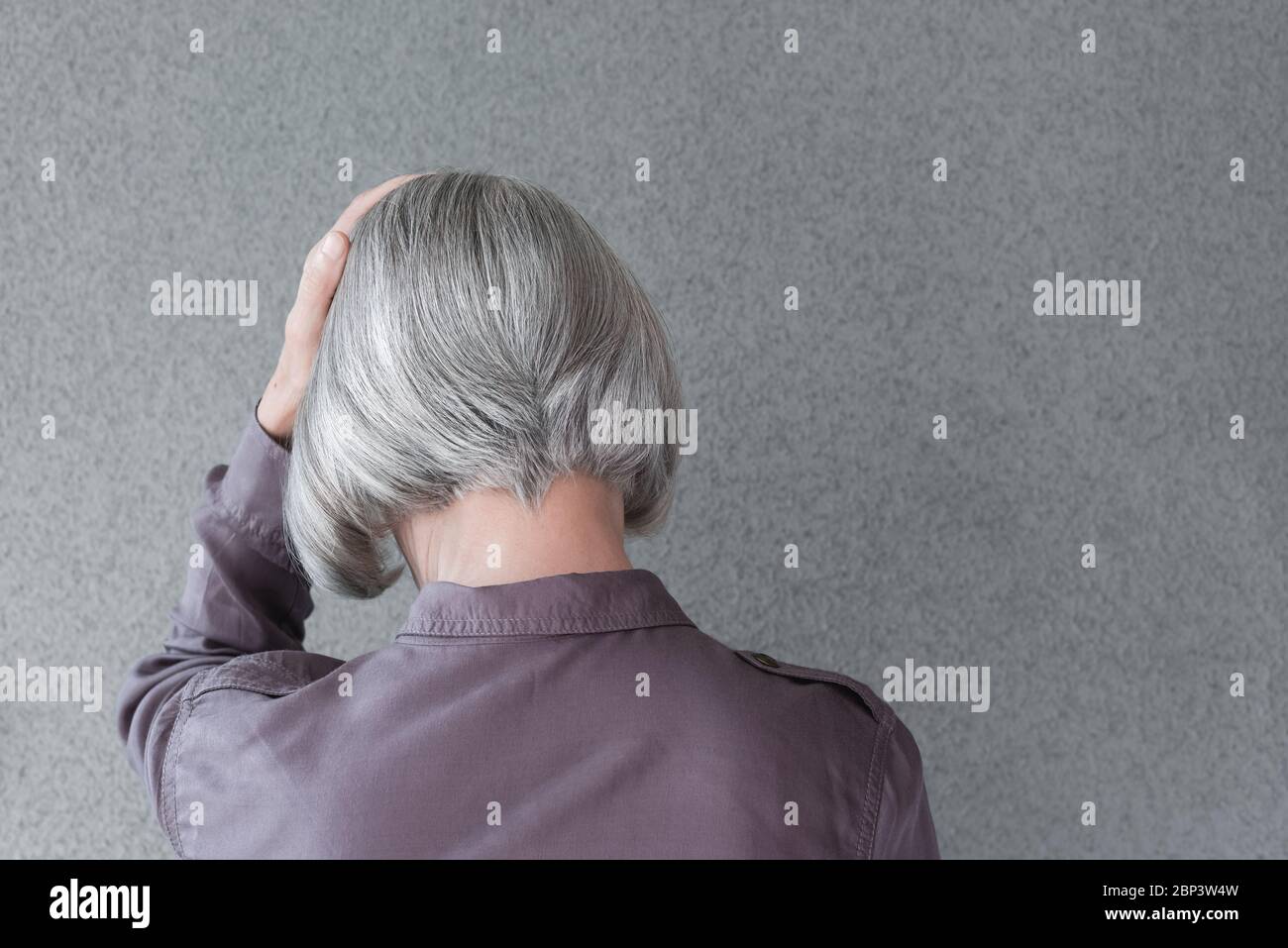 Upset gray-haired woman, holding her head with her hand, on gray background with copy space. Stock Photo