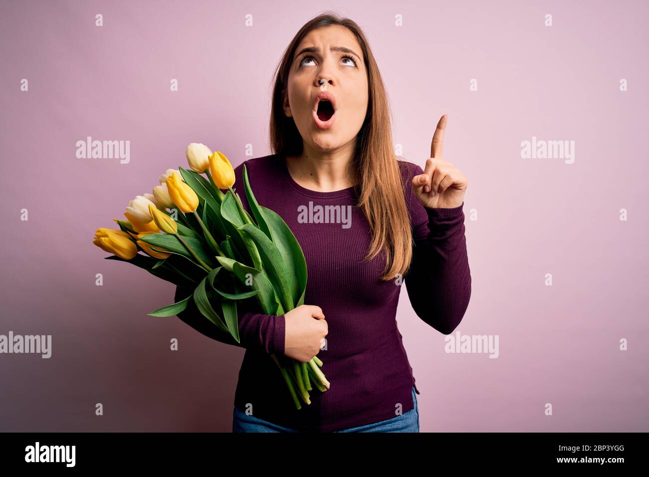 Young blonde woman holding romantic bouquet of yellow tulips flowers over pink background amazed and surprised looking up and pointing with fingers an Stock Photo