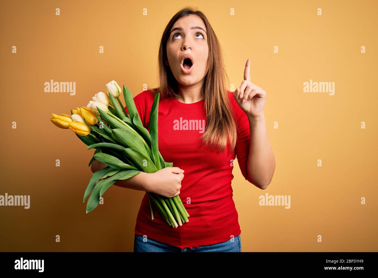 Young blonde woman holding romantic bouquet of tulips flowers over yellow background amazed and surprised looking up and pointing with fingers and rai Stock Photo