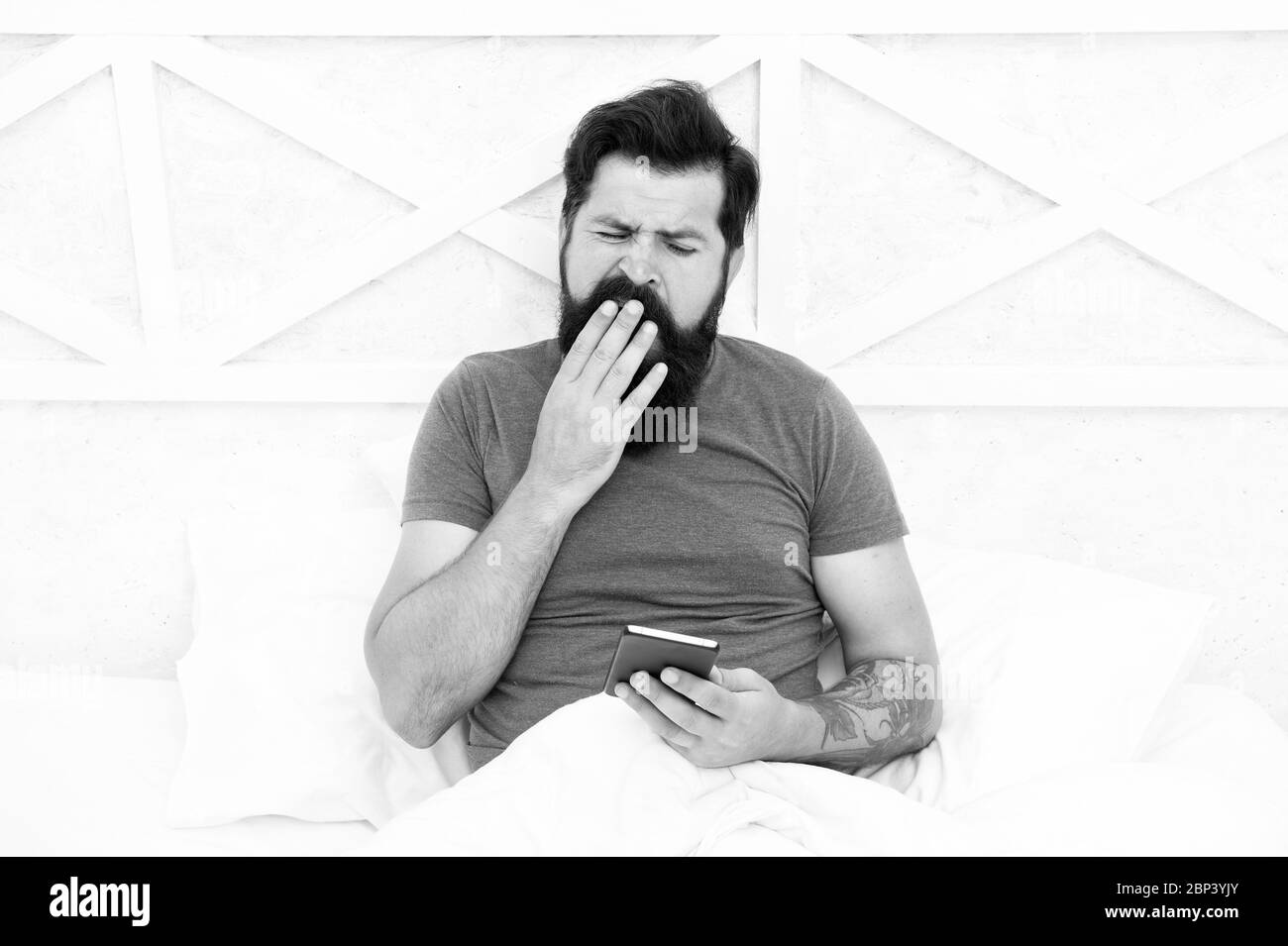 It takes him long to wake up. Sleepy man. Bearded man use mobile phone in bed. Man yawn in morning. Unshaven man with beard and mustache hair. Mobile lifestyle. Waking up. Good morning. Stock Photo