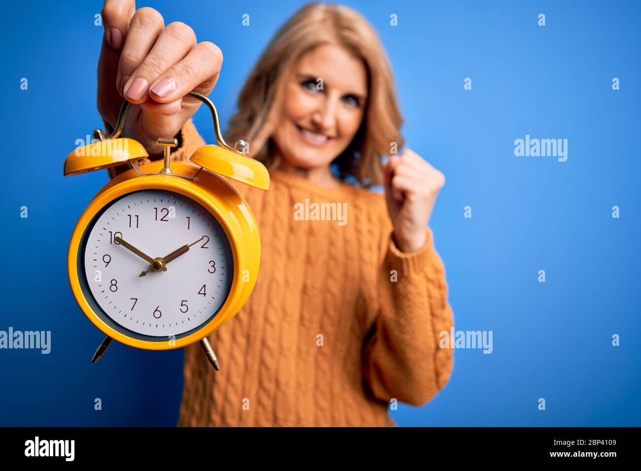 Middle age beautiful blonde woman holding vintage alarm clock over blue ...