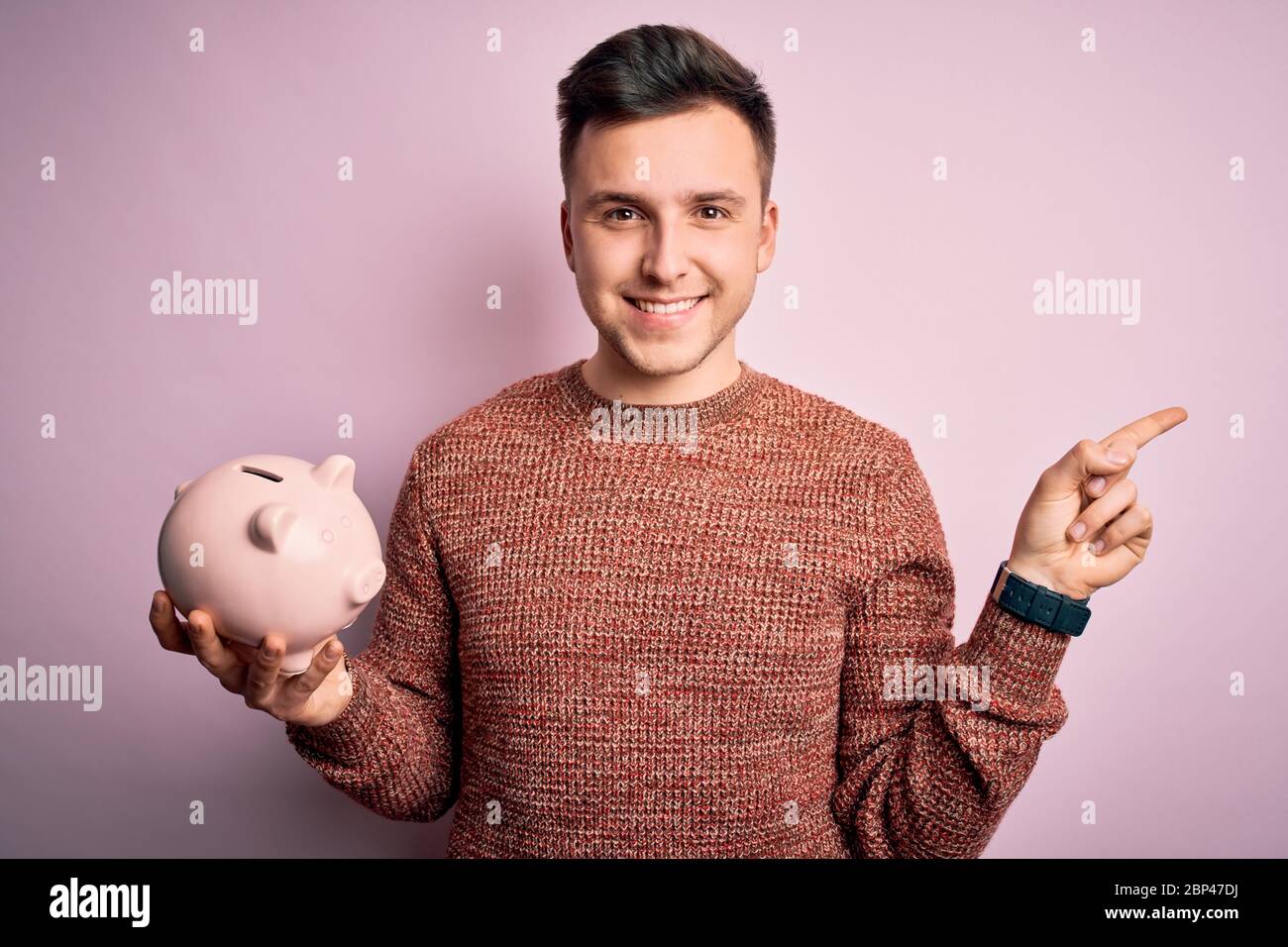Young handsome caucasian man holding piggy bank for savings over pink ...