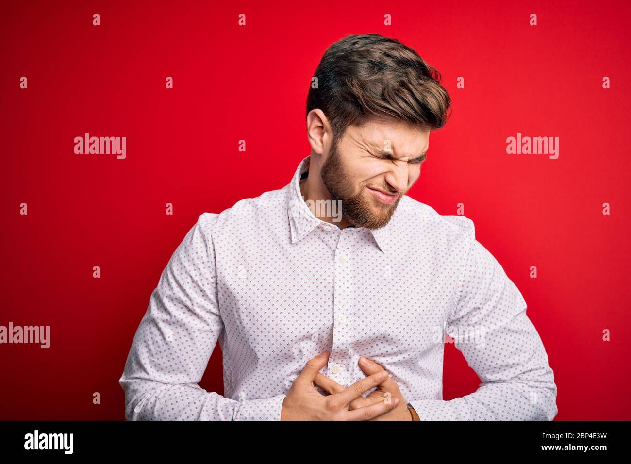 Young blond businessman with beard and blue eyes wearing elegant shirt over red background with hand on stomach because nausea, painful disease feelin Stock Photo