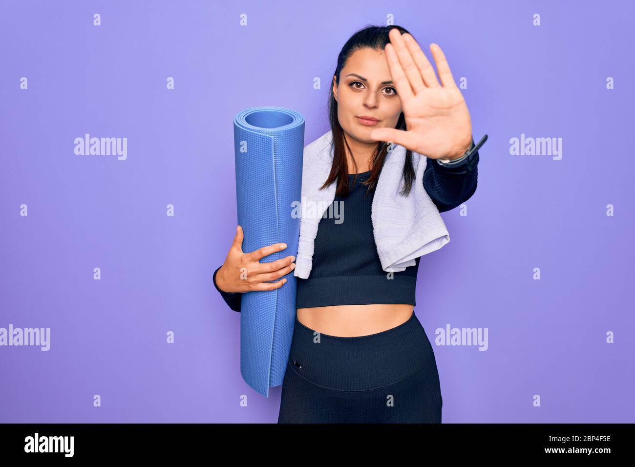 Young beautiful brunette sporty woman using sport towel hoding mat to do yoga with open hand doing stop sign with serious and confident expression, de Stock Photo