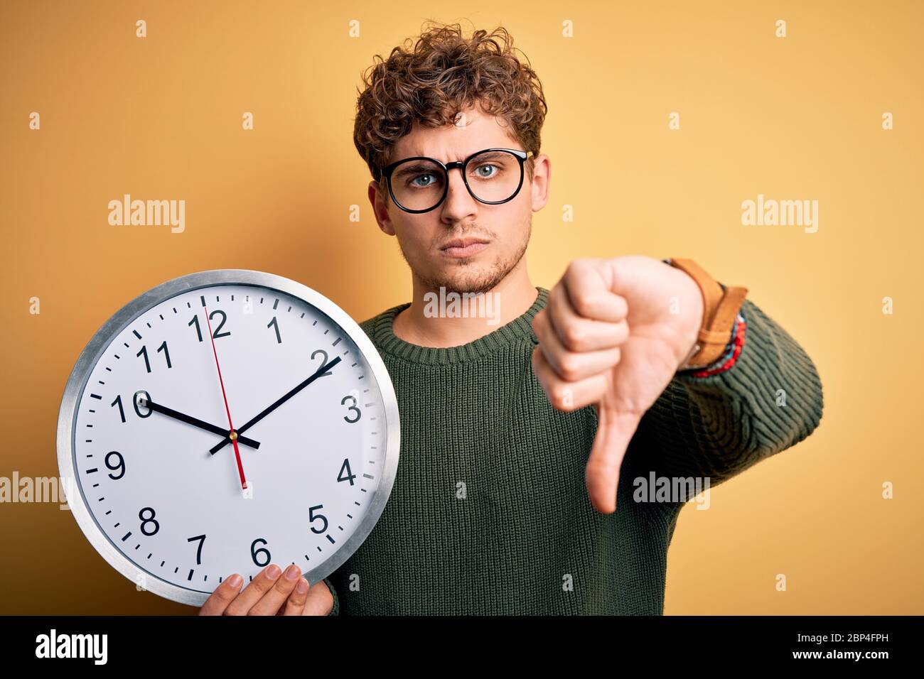 Young blond man with curly hair wearing glasses holding big clock over ...
