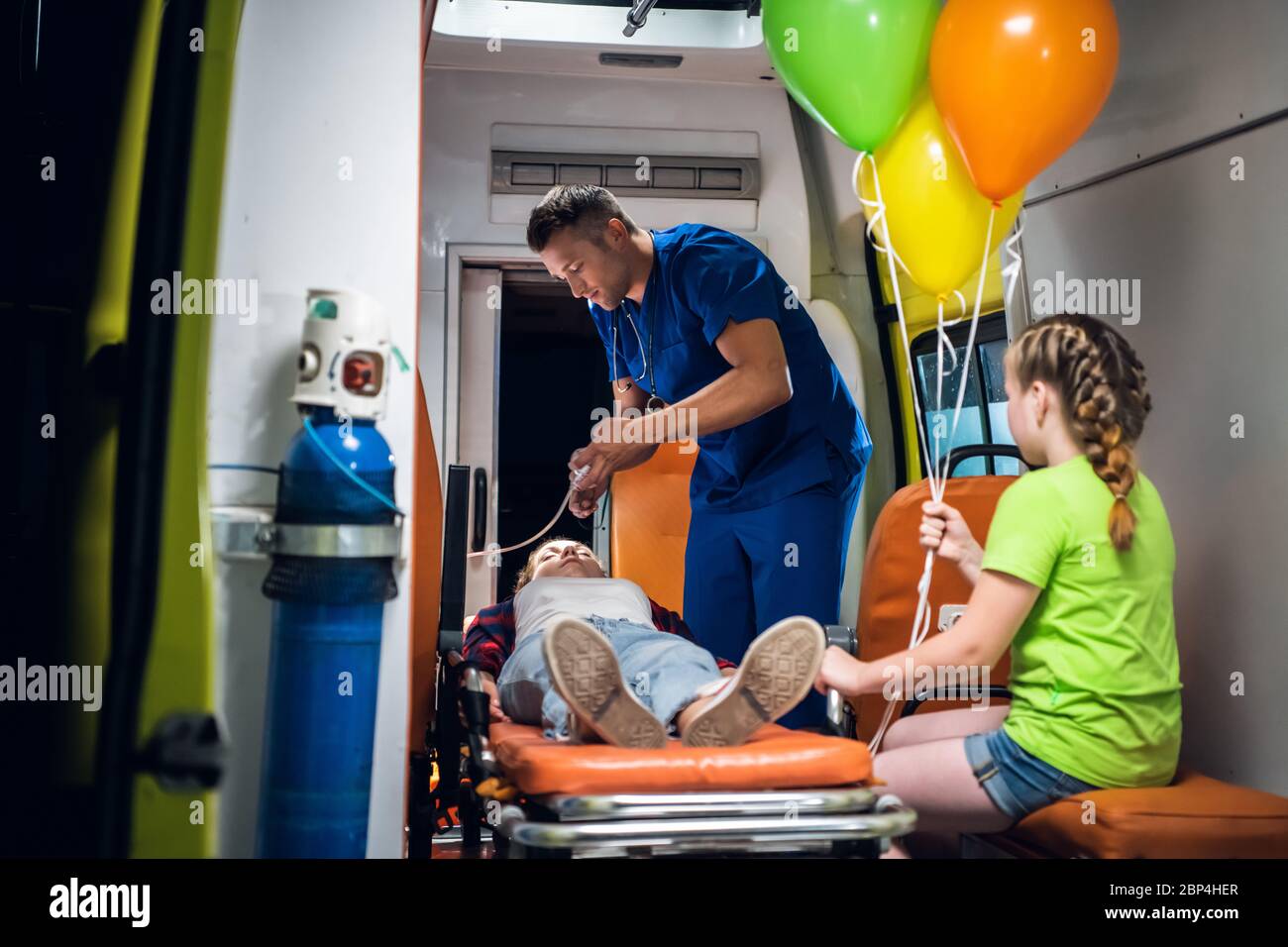 Man in a medical uniform puts an oxygen mask on a girl lying on a stretcher, her little sister is watching the doctor work Stock Photo