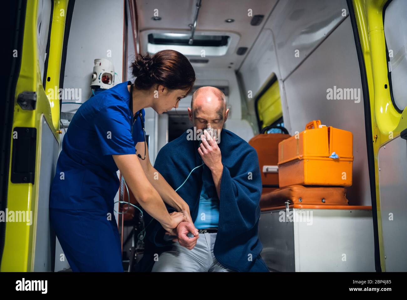 An injured, shocked man sitting with an oxygen mask in an ambulance, woman in a medical uniform is checking his pulse Stock Photo