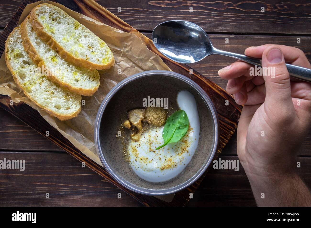 Man's hand holding spoon over a cappuccino soup with mushrooms on wooden cutting board. Three slices of white bread on the brown wooden background in Stock Photo