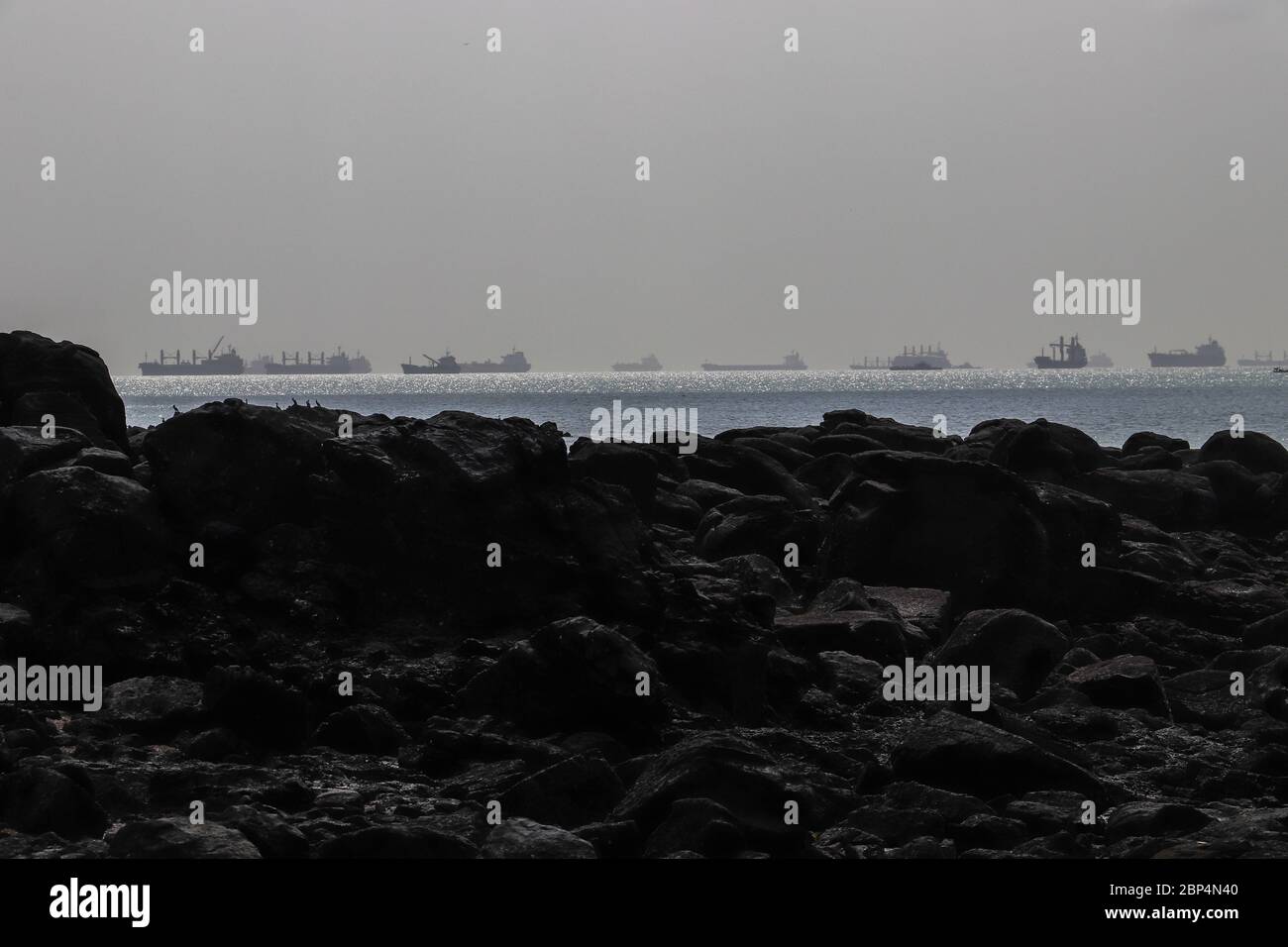 Huge ocean-going ships lined up on the horizon in a sparkling sea awaiting their turn to enter the Panama Canal as seen from a rocky shore. Copy space Stock Photo