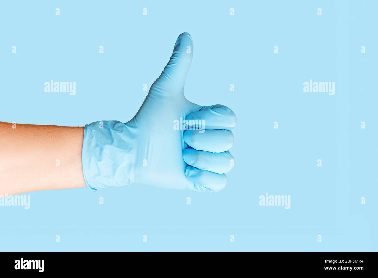 Hand wearing blue latex glove giving thumbs up on blue background. Stock Photo