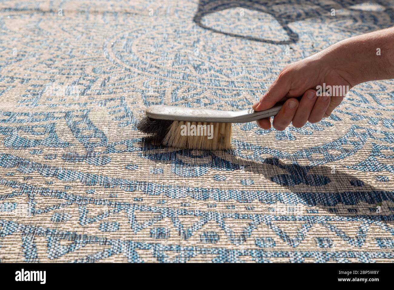Man brushing an outdoor rug in a garden with a hand brush. Grey and