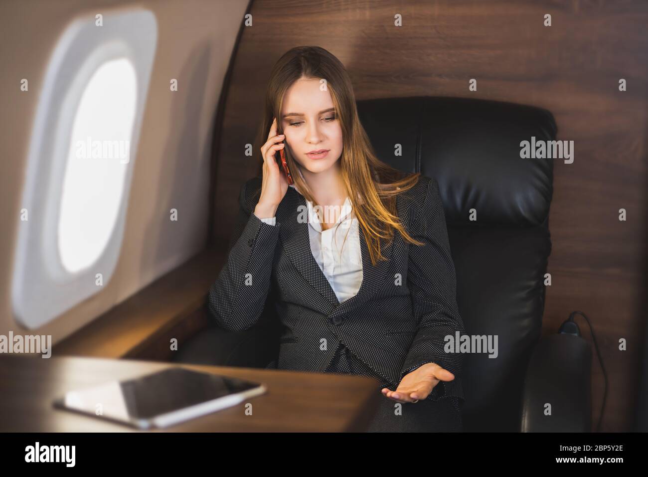 Attractive nervous young caucasian businesswoman is talking on the phone with an irritated facial expression, gesturing, sitting on a private plane. B Stock Photo