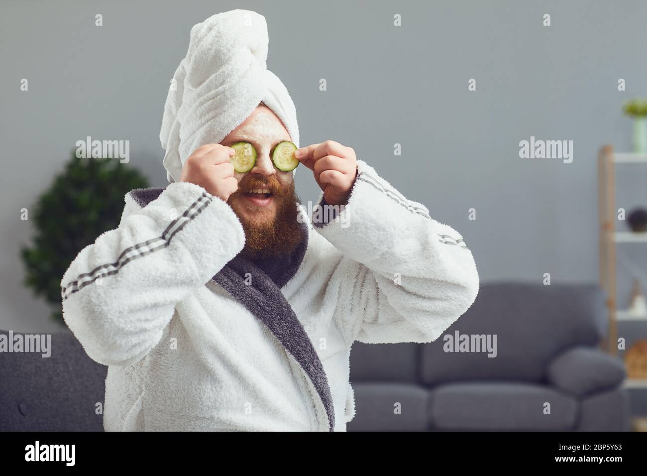Funny strange fat bearded man with a cosmetic mask on his face in a bathrobe does spa treatments on his face rests in a room at home Stock Photo