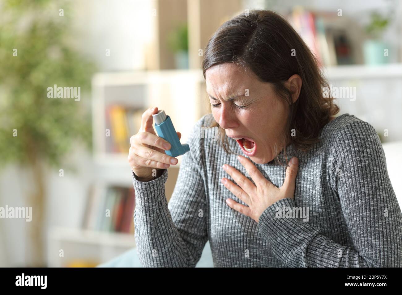 Asthmatic adult woman suffers asthma attack holding inhaler sitting on the couch at home Stock Photo