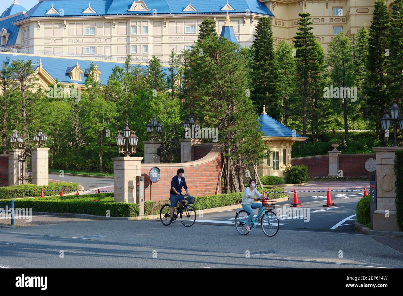 Cyclists wearing face masks pass a closed entrance to a Tokyo Disney Resort. Disneyland is closed and the area unusually quiet due to the coronavirus. Stock Photo