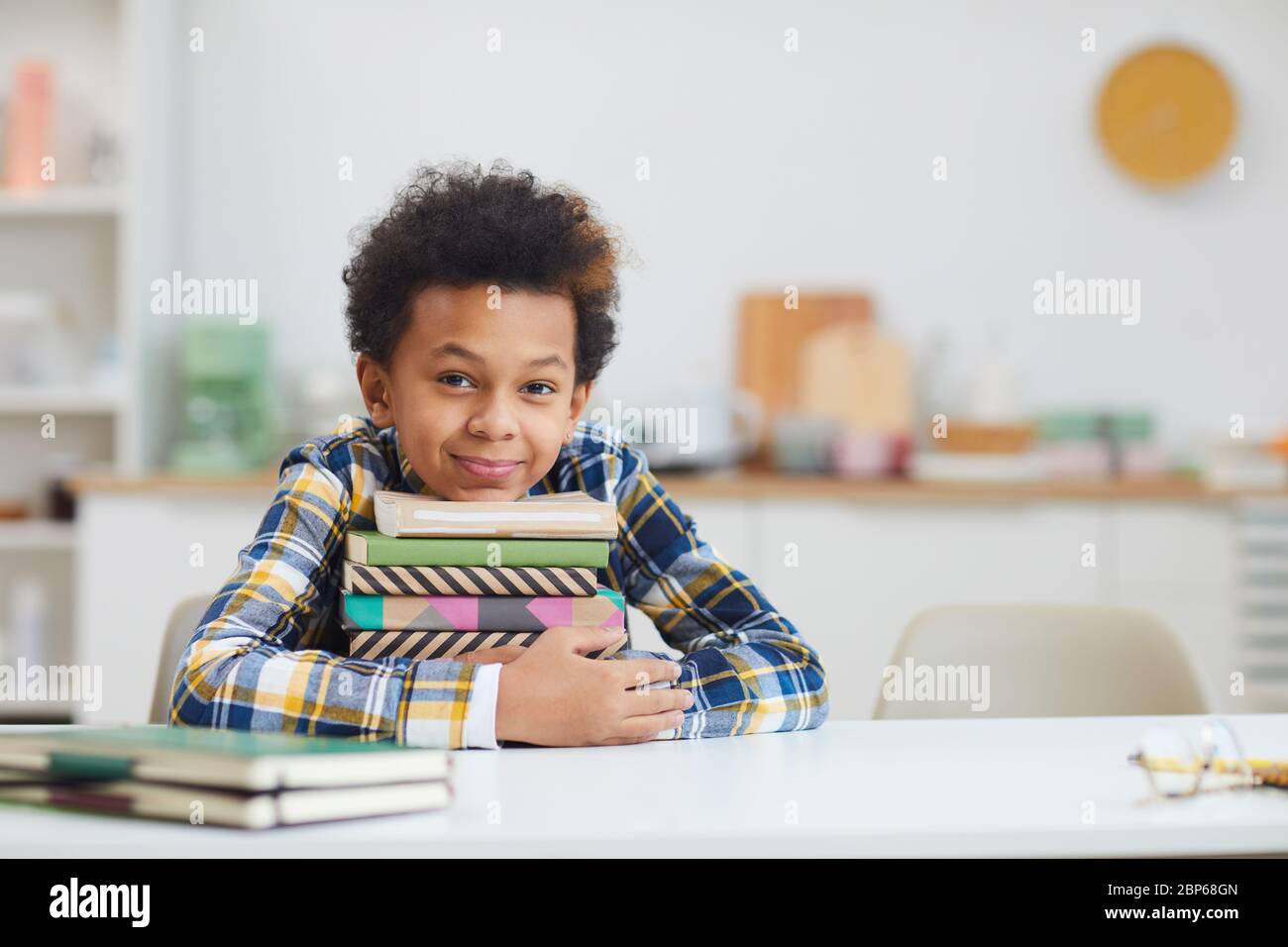 Portrait of cute African-American boy holding stack of books while sitting at desk at home and smiling at camera, copy space Stock Photo
