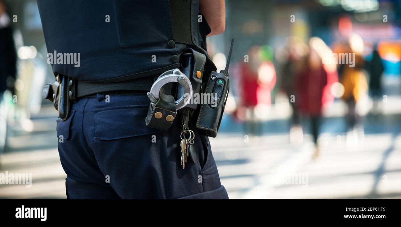 Police officer watching people. Detail Policeman with handcuffs and group of blurred people in background. Stock Photo