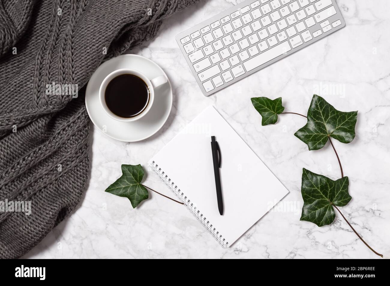 Cozy home office workspace. Marble desk with warm wool plaid, green ivy leaves, coffee cup, keyboard, blank paper in notebook and pen in black and whi Stock Photo