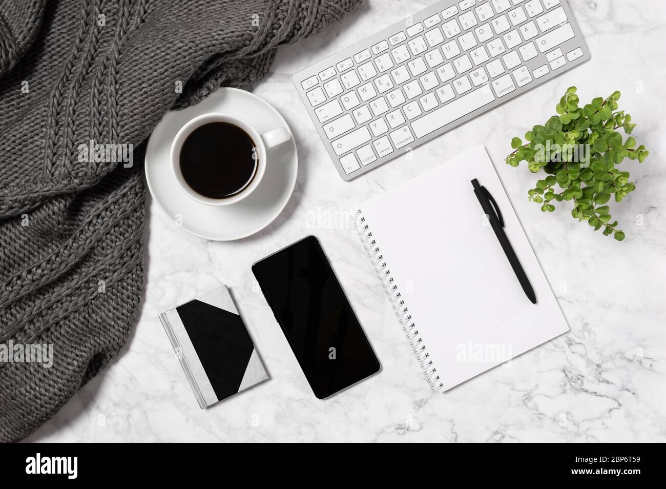 Cozy home office workspace. Marble desk with warm wool plaid, cup of coffee, keyboard, mobile phone, green house plant, blank paper, notebook and pen Stock Photo