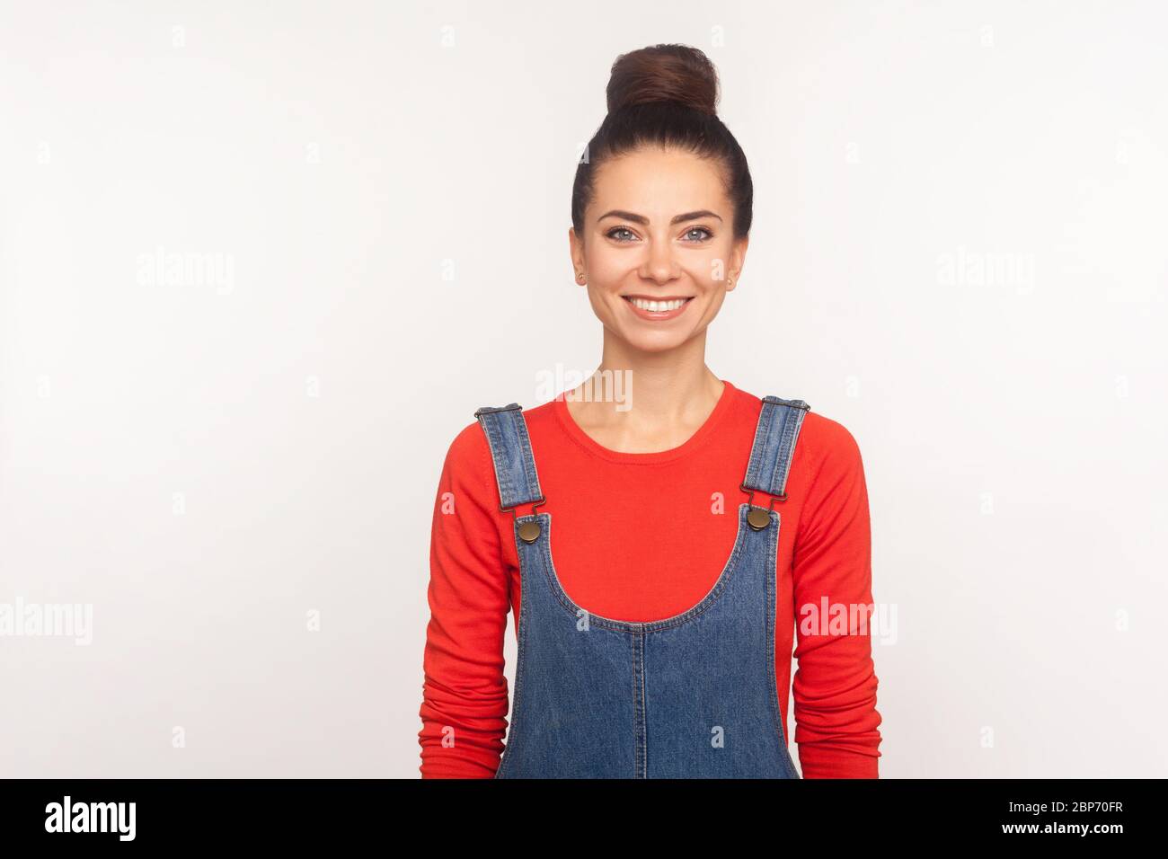 Portrait of positive happy stylish pretty girl with hair bun in denim overalls looking at camera with engaging toothy smile, expressing joy and optimi Stock Photo