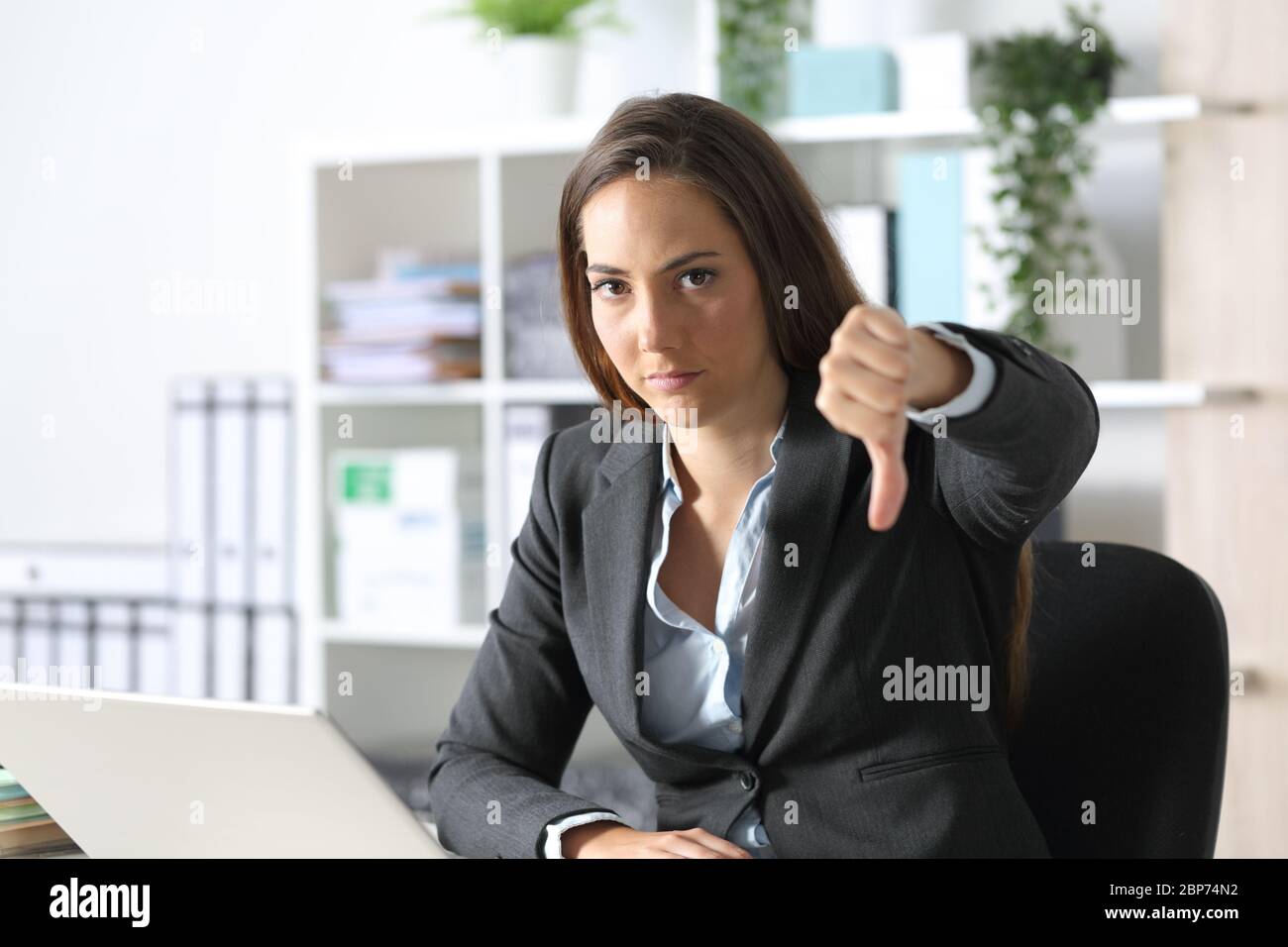 Serious executive woman gesturing thumbs down looking camera sitting on a desk at the office Stock Photo