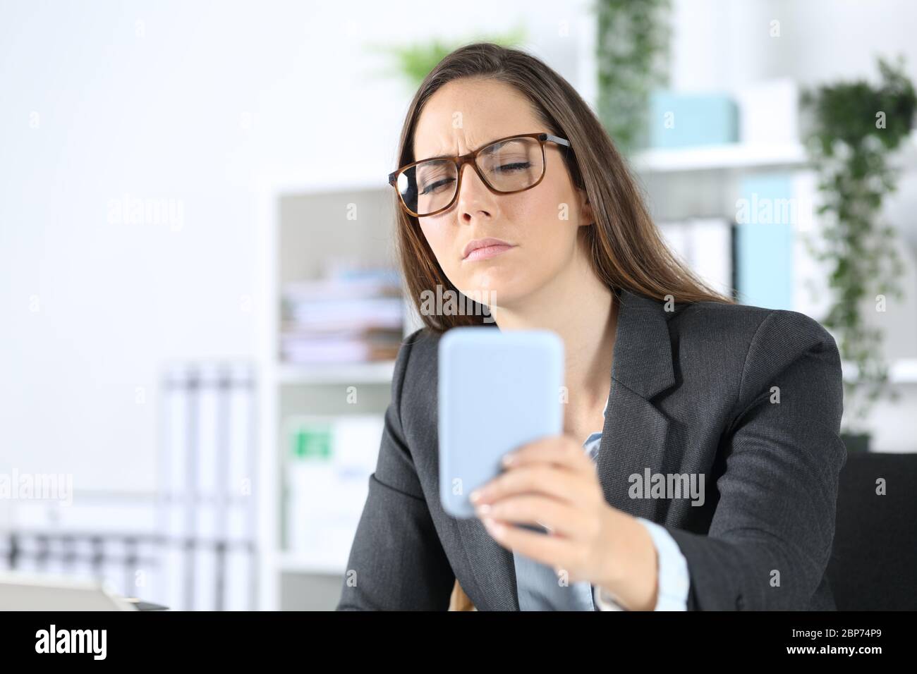 Executive woman wearing eyeglasses with eyesight problem trying to read on smart phone sitting on her desk at office Stock Photo