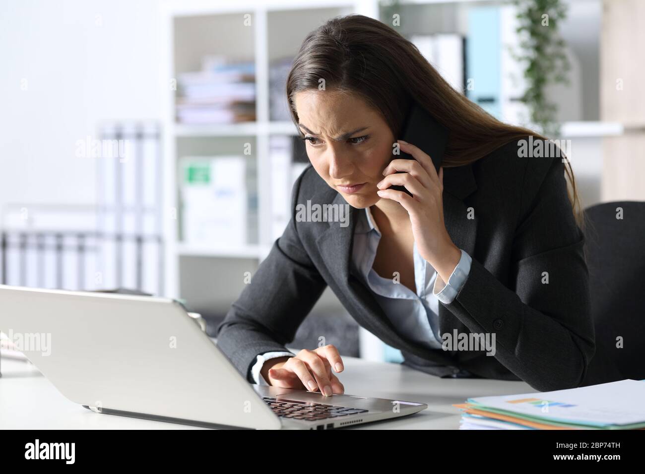 Suspicious executive woman calling on smart phone looking at laptop sitting on her desk at office Stock Photo