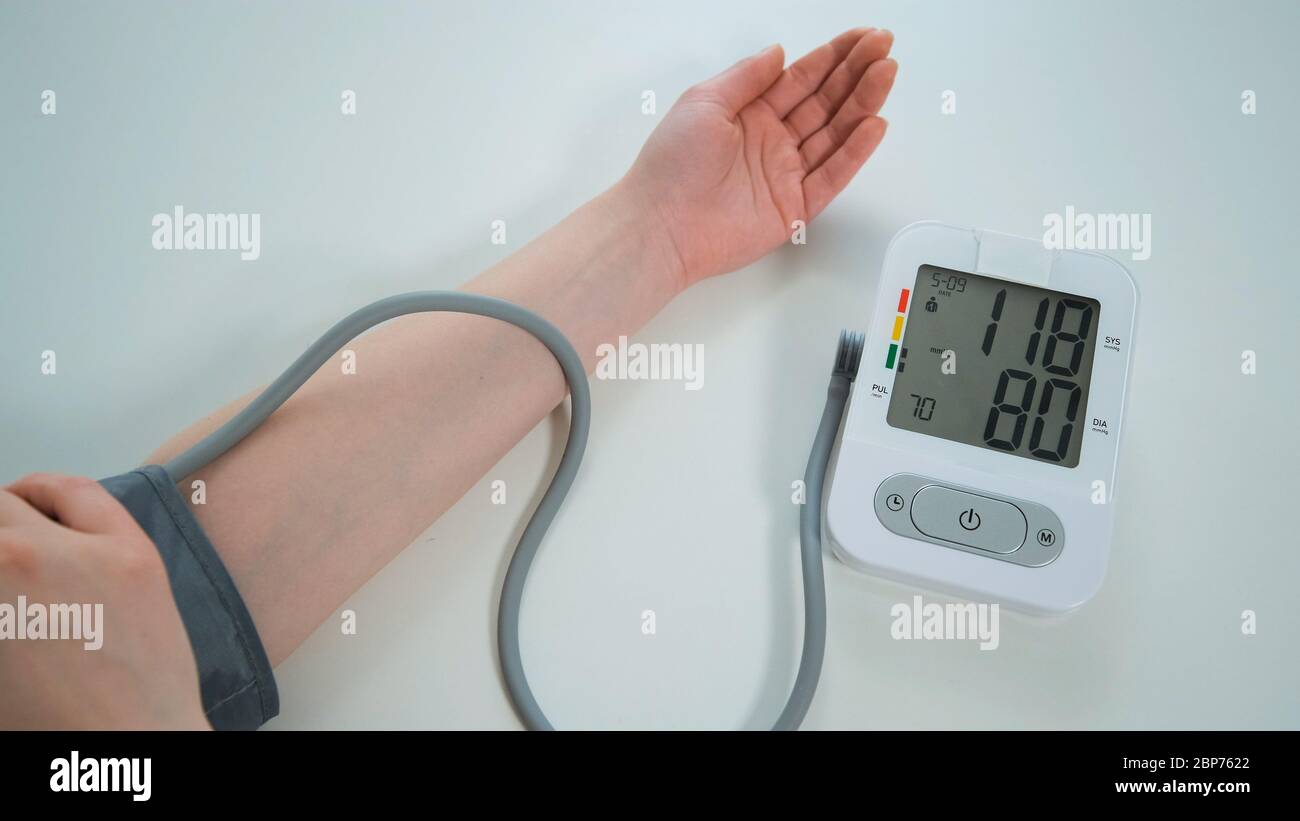 Woman measures his blood pressure at the table. Modern automatic white and grey tonometer displaying on monitor the numbers of measurement. Stock Photo