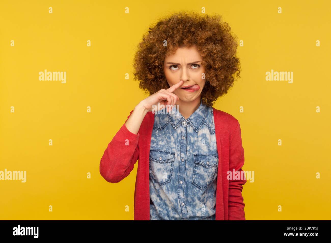 Portrait of funny woman with fluffy curly hair drilling nose fooling around, showing tongue out and picking nose with comical silly expression. indoor Stock Photo