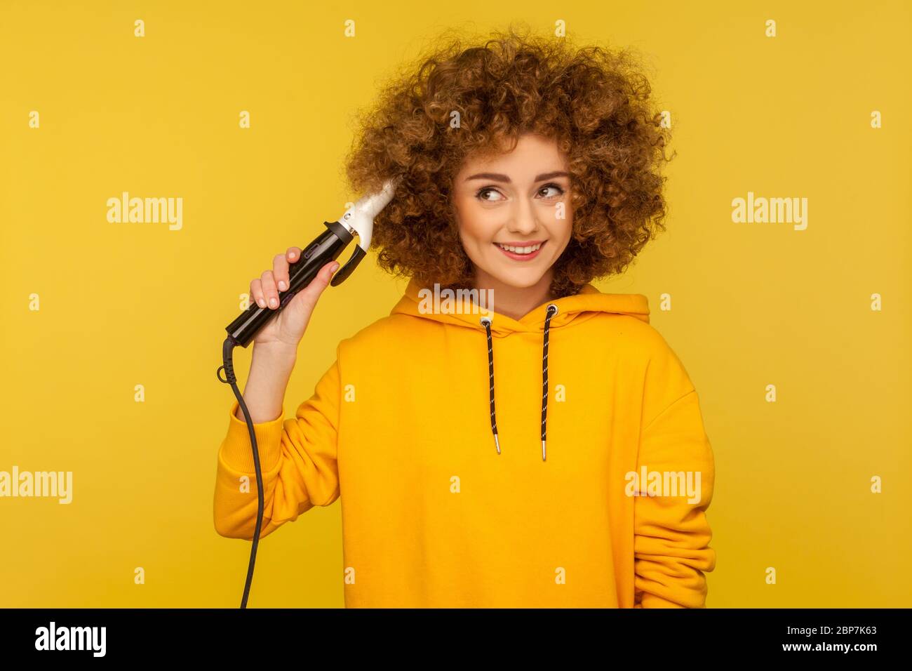 Fancy positive woman holding curling iron, making perfect afro hairdo, trendy girl ironing hair curls, grooming with professional tool, beauty care co Stock Photo
