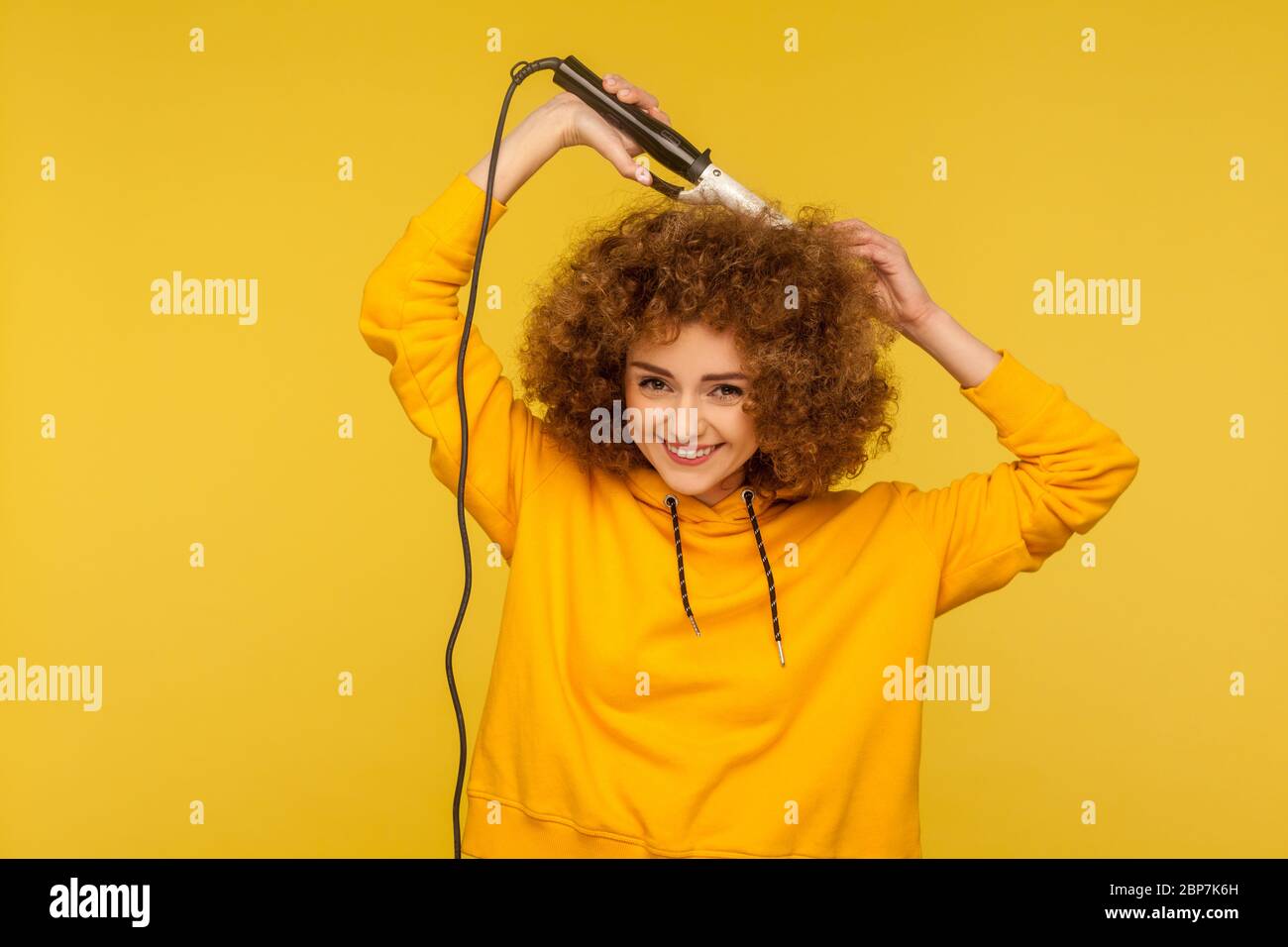 Fancy cheerful woman using curling iron for perfect afro hairdo, trendy girl ironing hair curls, grooming with professional tool, beauty care concept. Stock Photo