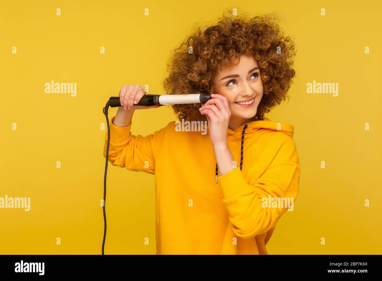 Beautiful happy woman using curling iron for perfect afro hairdo, trendy girl ironing hair curls, grooming with professional tool, beauty care concept Stock Photo