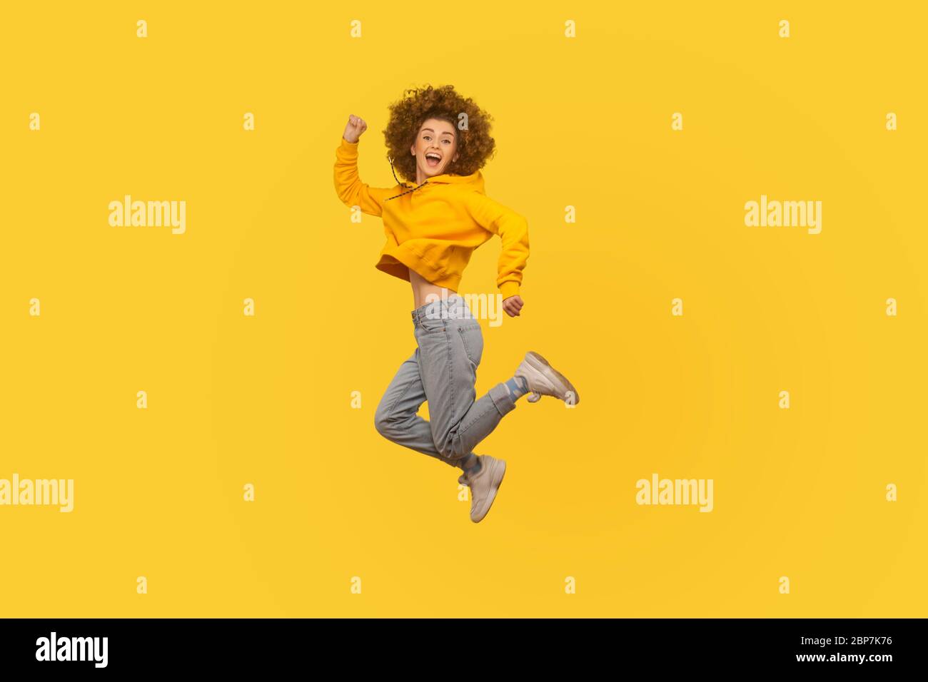 Portrait of enthusiastic joyful curly-haired girl in urban style hoodie and jeans jumping high in air, flying and shouting with amazed happy expressio Stock Photo