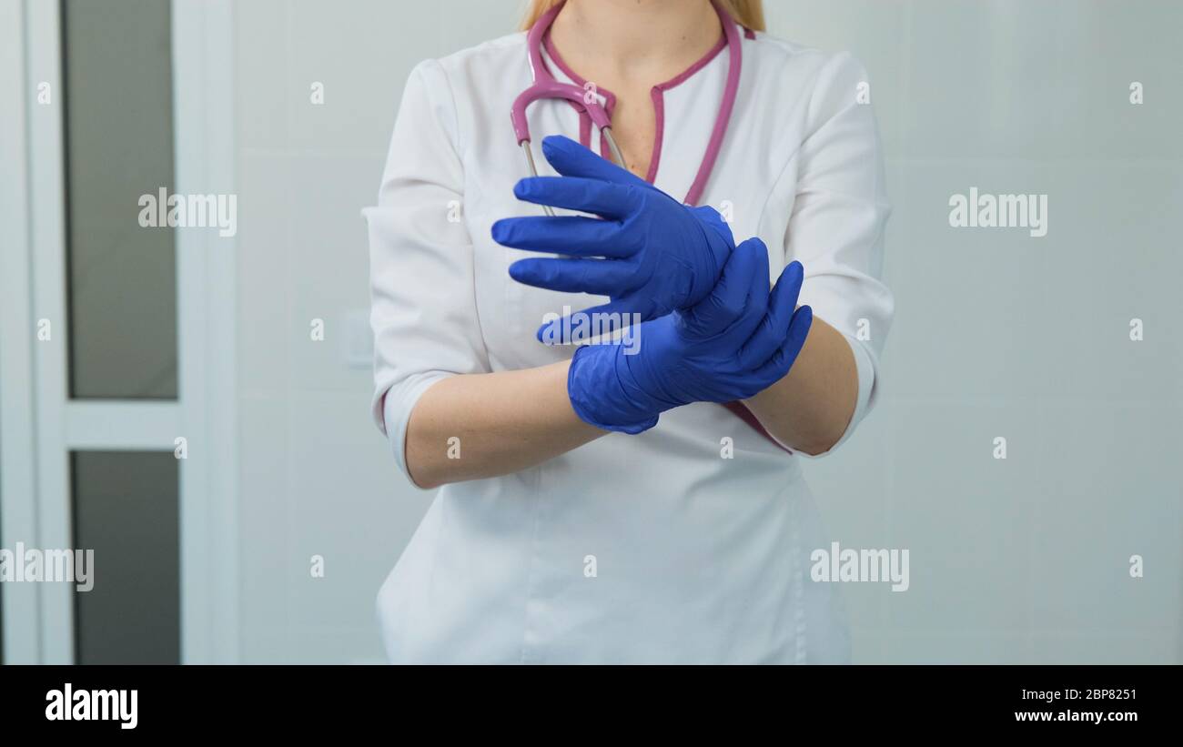 Handsome young women doctor in white medical coat, violet stethoscope putting on rubber blue gloves in hospital or clinic. Front view on a white backg Stock Photo