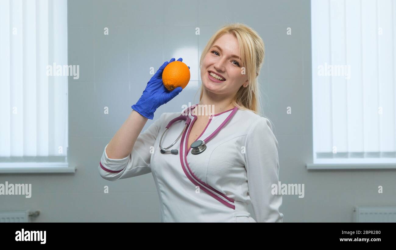 Caucasian young blonde doctor in white medical coat, violet stethoscope holding a white pills with left hand and orange in rigth hand in blue gloves a Stock Photo